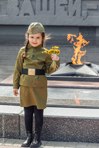 Patriotic education of children. Little girl with long braids in a military uniform, military cap, holds flowers in hands against the background of the memorial. Photo for the Victory Day on May 9