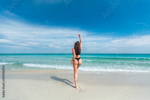 Fototapeta Naklejka Na Ścianę i Meble -  Beautiful young girl in the black swimsuit and sunglasses is smiling and standing on the beach on the background of azure caribbean sea.