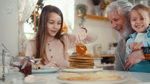Joyful grandfather and his grandchildren at the table enjoy eating pancakes in the kitchen, cute girl pours honey over pancakes while little boy and grandpa laugh and have fun.