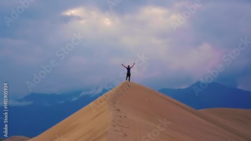 Person standing on sand dune with arms raised under dramatic sky