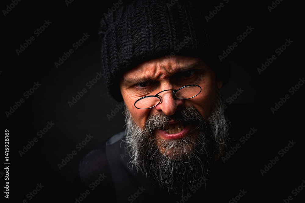 Stylish portrait of a confident mature man with his arms crossed in front of him, wearing a knitted hat and glasses. Staring, gray beard and black vest. Stylish discreet portrait on a dark background,