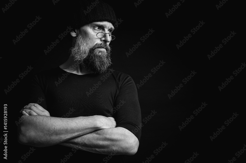 Black and white, stylish portrait of a confident mature man with his arms crossed in front of him, wearing a knitted hat and glasses. Staring, gray beard and black vest. Stylish discreet portrait