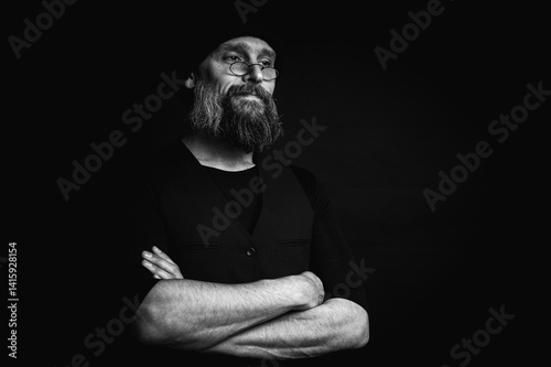 Obraz na płótnie Black and white, stylish portrait of a confident mature man with his arms crossed in front of him, wearing a knitted hat and glasses. Staring, gray beard and black vest. Stylish discreet portrait w salonie