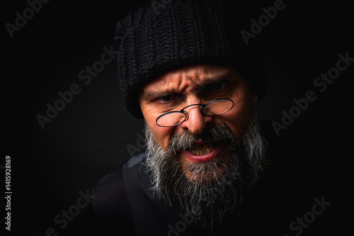 Obraz na płótnie Stylish portrait of a confident mature man with his arms crossed in front of him, wearing a knitted hat and glasses. Staring, gray beard and black vest. Stylish discreet portrait on a dark background, w salonie