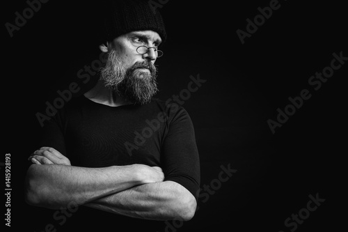Obraz na płótnie Black and white, stylish portrait of a confident mature man with his arms crossed in front of him, wearing a knitted hat and glasses. Staring, gray beard and black vest. Stylish discreet portrait w salonie