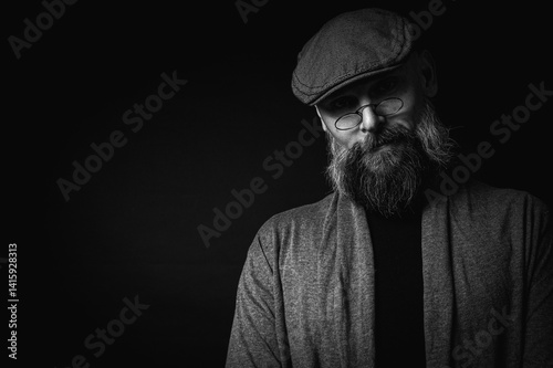 Obraz na płótnie Black and white, Macro portrait of a serious bearded man in a cap and glasses on a black background. Stylish, intense look with a confident and mysterious expression, piercing gaze w salonie