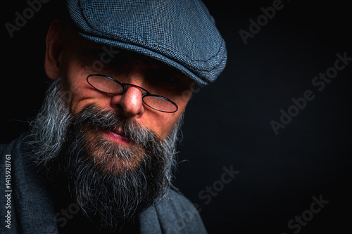 Close-up portrait of a serious bearded man in flat cap and glasses on black background. Stylish, intense look with confident and mysterious expression