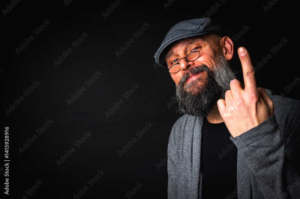 Elderly bearded man in cap and glasses shows a finger up gesture, expressing sarcasm or rebellion. Studio portrait on black background