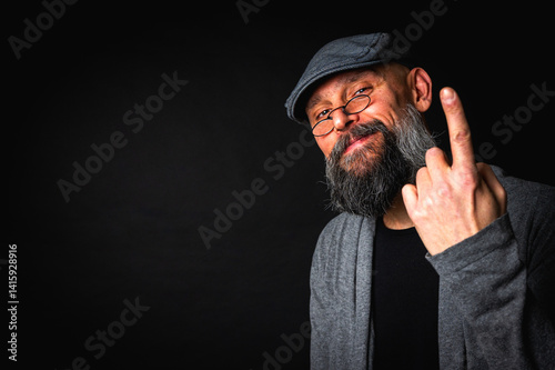 Obraz na płótnie Elderly bearded man in cap and glasses shows a finger up gesture, expressing sarcasm or rebellion. Studio portrait on black background w salonie