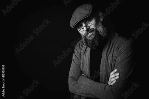 Black and white, Macro portrait of a serious bearded man in a cap and glasses on a black background. Stylish, intense look with a confident and mysterious expression, piercing gaze