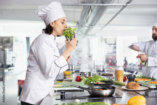 Obraz na płótnie Dedicated female chef smelling the aroma of herbs and greenery, using tasty fresh rosemary or parsley leaves and sprinkling in the pan. Culinary expert cooking dishes with raw materials, gastronomy. w salonie