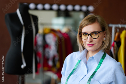 confident fashion designer in glasses with measuring tape around neck in creative studio. professional workspace with dress form and vibrant fabrics in background.