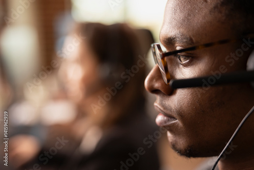 Closeup of african american man with glasses, wearing a headset with microphone while speaking with online customers. Focused male telemarketer working in startup office, providing technical support.