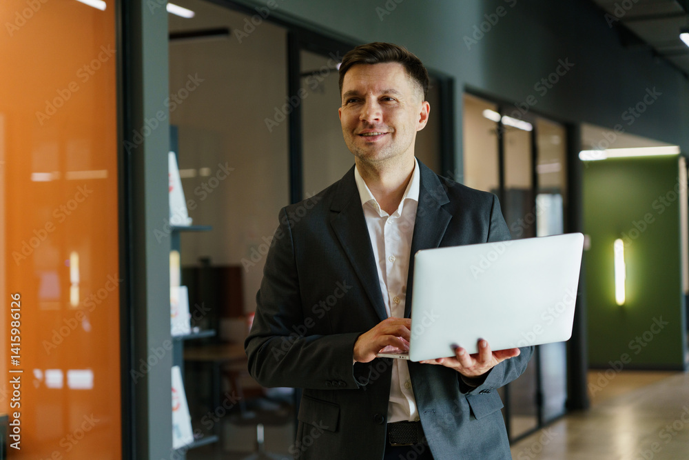 Professional Man Smiles Confidently While Using a Laptop in a Modern Office Corridor During the Day Professional Man Smiles Confidently While Using a Laptop in a Modern Office Corridor During the Day