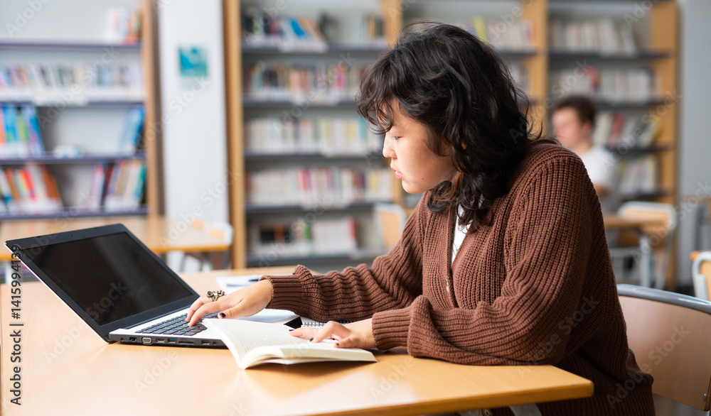 Asian fifteen-old-year schoolgirl searches for information in a book and types on a laptop while doing homework in the school library