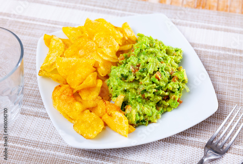 On table in cafe there is plate with traditional Mexican snack guacamole and pile of crispy potato chips. Salty and savory light repast for beer