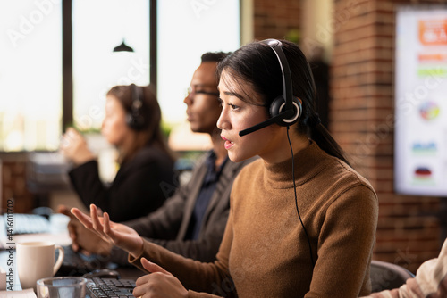 Obraz na płótnie Asian businesswoman wearing wired headphones and providing assistance to remote clients. Serious female call operator with headset sitting at desk and handling customer inquiries. w salonie