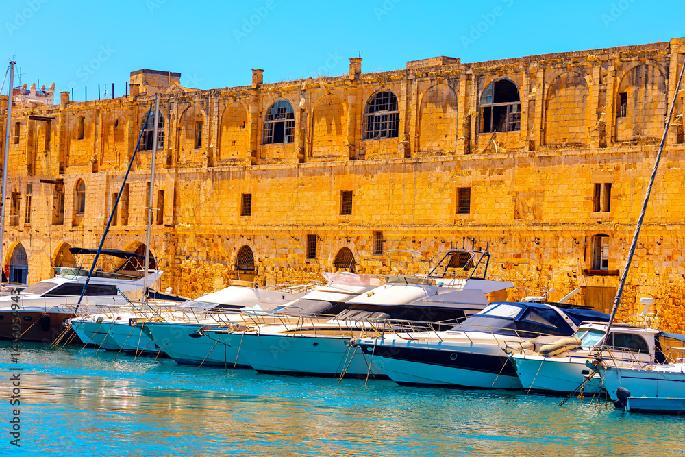 Row of modern yachts are docked along historic stone fortress with arched windows in Malta, under blue sky. Scene captures island blend of ancient architecture and contemporary luxury by waterfront