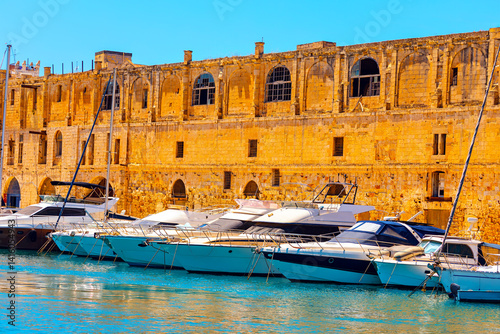 Obraz na płótnie Row of modern yachts are docked along historic stone fortress with arched windows in Malta, under blue sky. Scene captures island blend of ancient architecture and contemporary luxury by waterfront w salonie