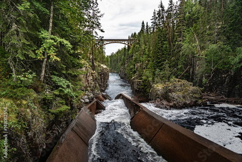 A railway bridge arches over the deep canyon of Storstupet, with rushing waters of the River Ämån below, surrounded by dense trees a log flume in a rugged terrain in Orsa Dalarna