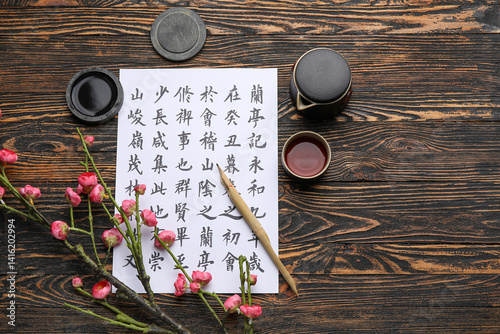 Tablou pe pânză Paper sheet with Asian hieroglyphs, nib pen, ink, cup of tea and blooming branch on wooden background