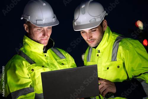 Engineer worker watching work monitor on laptop and checking for quality control at construction site of petroleumchemical industry or refinery oil plant factory overtime at night