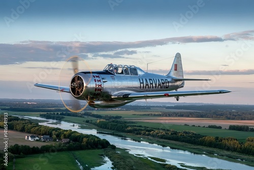 Vintage silver harvard aircraft flying above a river with green fields and a cloudy sky view