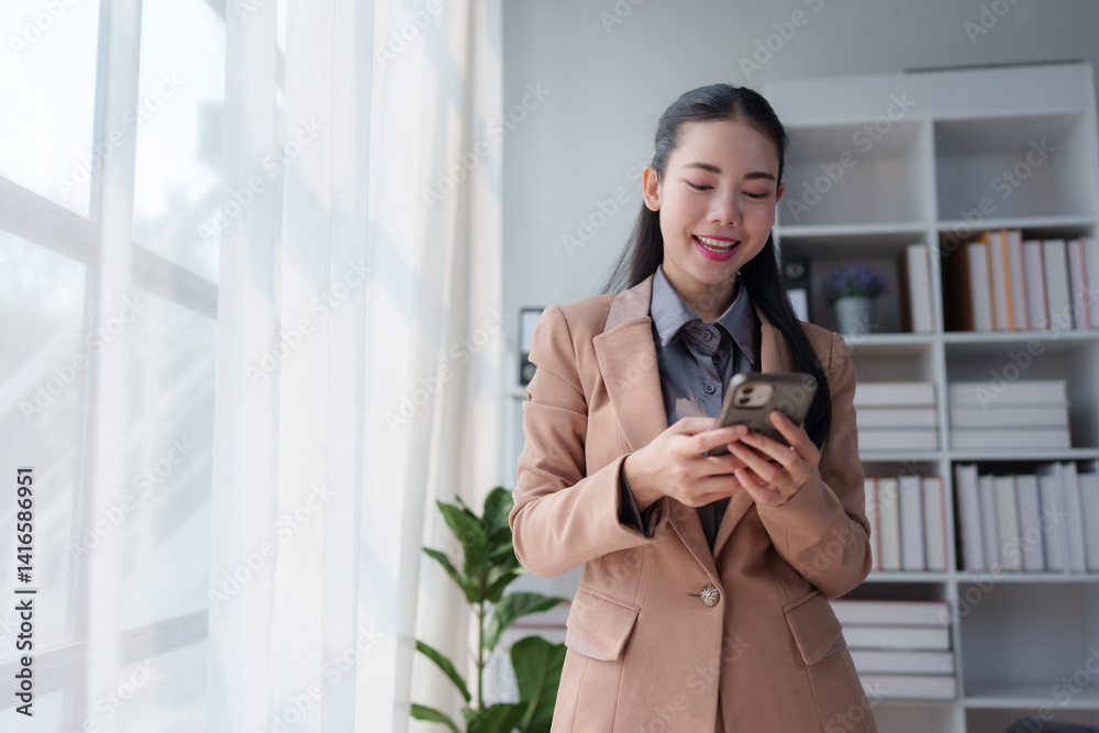Fototapeta premium Young asian manager wearing a tailored suit, using a mobile phone in a modern office filled with bookshelves and natural light, smiling while checking messages on the screen