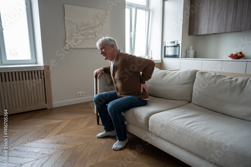 Abandoned senior man clutches painful spine unable to stand from couch due to multiple health issues. Herniated disc agony, pinched nerves, chronic kidney disease, mobility loss, self care inability