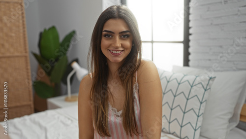Woman smiling on bed in bedroom with natural lighting, featuring brunette hair, hispanic descent, and casual attire in a cozy, stylish home interior setting.