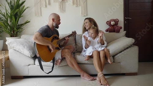 Father plays guitar and singing for his family, daughter and mother listening and applaud. Home activity for dad, mom and child. 