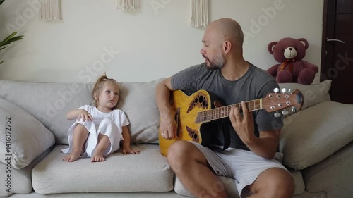 Father plays the guitar and singing for his small daughter sitting on sofa. Cozy family of dad and his child in a modern home, activity, parenting. Happy memories of childhood and fatherhood