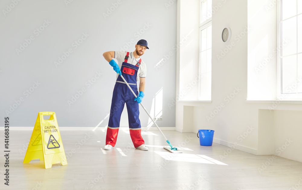 Male young bearded janitor in uniform washing floor with caution wet floor sign in empty room or at office. Man cleaner with mop and bucket mopping indoors. Cleaning service concept.