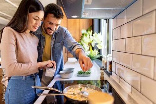 Young loving couple cooking together adding spices and salt in a pan with pasta in a modern kitchen