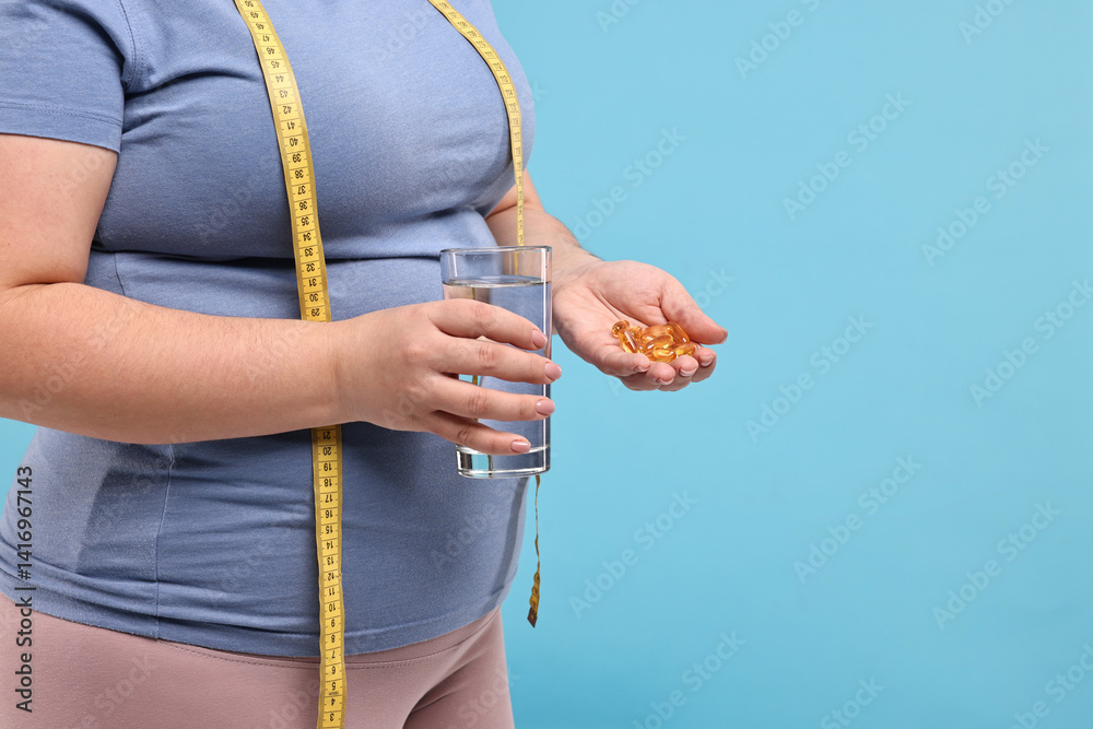 Plus size woman holding pile of weight loss supplements and glass with water on light blue background, closeup. Space for text