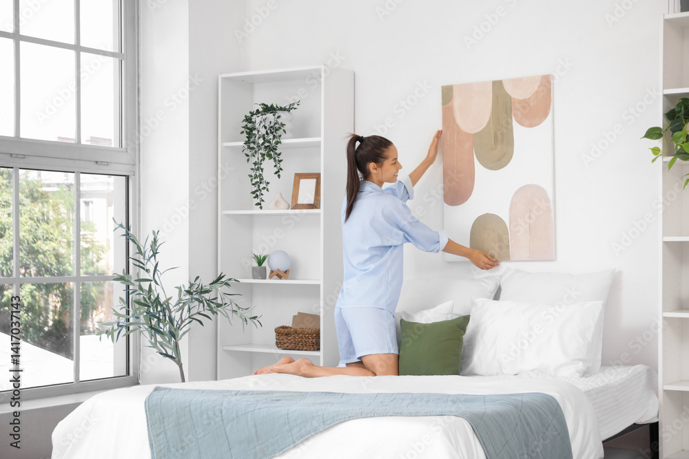 Young woman hanging stylish painting on light wall in bedroom