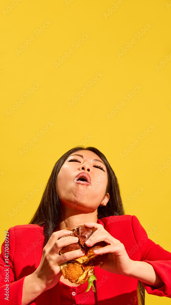 Vertical portrait of young Asian woman eating burger and expressing satisfaction of its delicious taste against bright yellow background. Concept of food, pop art, human emotions, delight