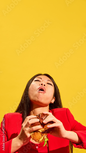 Obraz na płótnie Vertical portrait of young Asian woman eating burger and expressing satisfaction of its delicious taste against bright yellow background. Concept of food, pop art, human emotions, delight w salonie