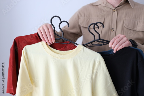 A person is holding up three shirts on hangers, one of which is yellow. The person is wearing a brown shirt and a watch. The shirts are hanging on a rack