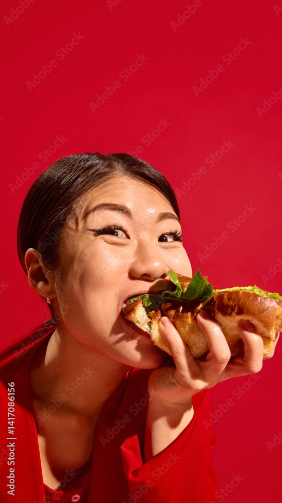 Happy Asian woman biting delicious sandwich with joy, wide-eyed and expressive face against bold red background. Concept of food culture, pop art photography, human emotions. Vertical layout