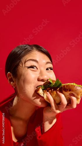 Obraz na płótnie Happy Asian woman biting delicious sandwich with joy, wide-eyed and expressive face against bold red background. Concept of food culture, pop art photography, human emotions. Vertical layout w salonie