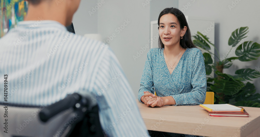 Fototapeta premium In a professional office setting, a wheelchair user in a shirt sits across from a beautiful Asian-Korean woman during a job interview. She listens carefully, taking notes as he answers her questions.