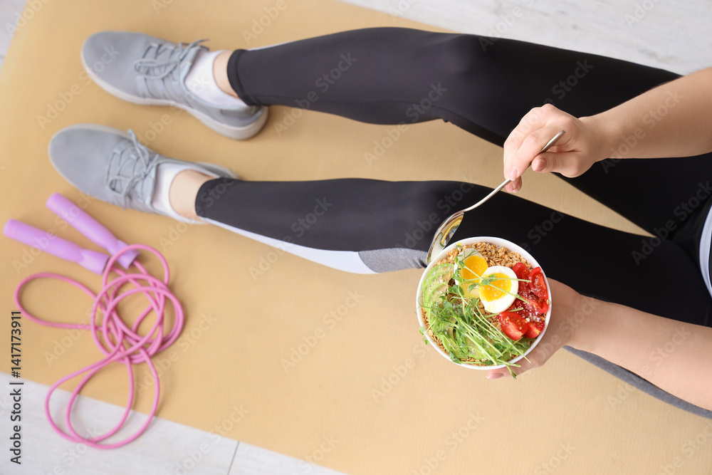 Sporty young woman sitting on yoga mat with bowl of nutritious oatmeal and skipping rope at home, top view