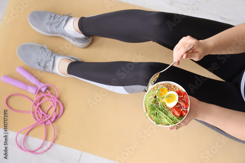 Sporty young woman sitting on yoga mat with bowl of nutritious oatmeal and skipping rope at home, top view