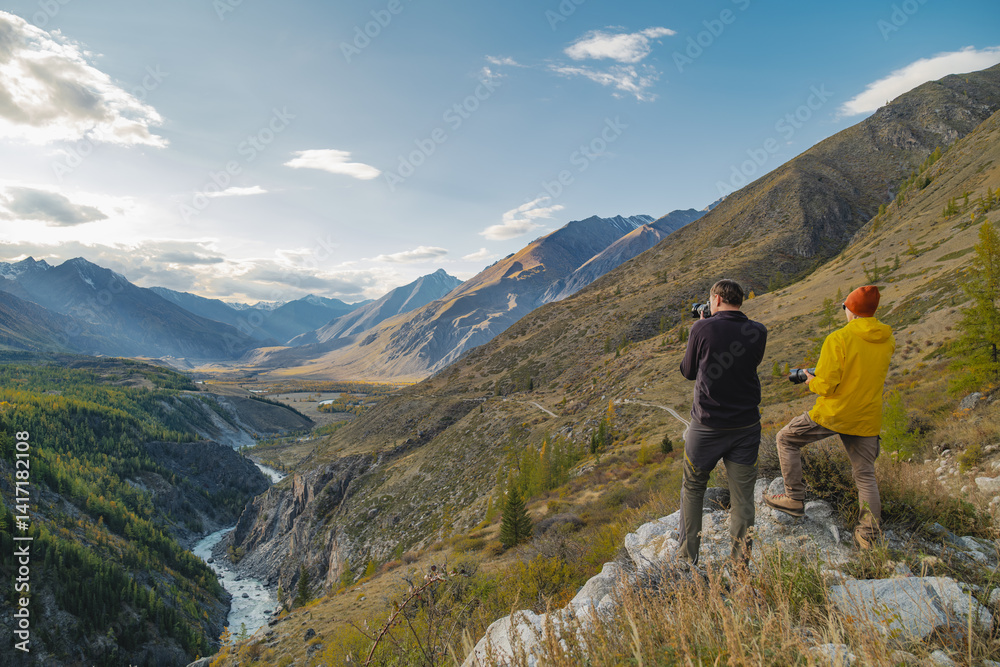Fototapeta premium Two photographers with cameras are standing on a slope in bright clothes and taking pictures against the background of snowy mountains, forests and a river that flows through the Altai gorge.