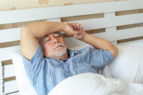 Asian senior man lying in bed with closed eyes and hand on forehead, suffering from insomnia, fatigue or headache, elderly person resting in bedroom, concept of health problems and aging
