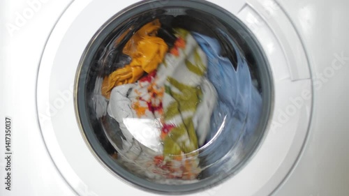 Rotating washing machine drum tumbling colorful wet garments, splashing water and detergent during laundry cleaning process