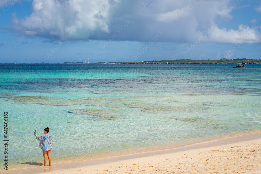 Woman takes selfie on Okinawa beach, capturing the turquoise water and sunny skies..