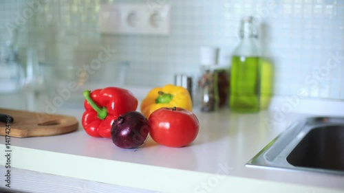 a woman placing a vegetables on a kitchen countertop, surrounded by fresh vegetables like tomatoes, cucumbers, bell peppers, and a purple onion. Olive oil is visible in the background.