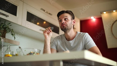 A man in a gray t-shirt is enjoying a bite of salad from a fork in a modern kitchen. He looks satisfied while eating, with a bowl of salad on the counter in front of him.